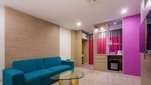 Una sala de estar moderna con un sofá azul verdoso, una mesa de centro de vidrio y una zona de kitchenette con cortinas rosas.