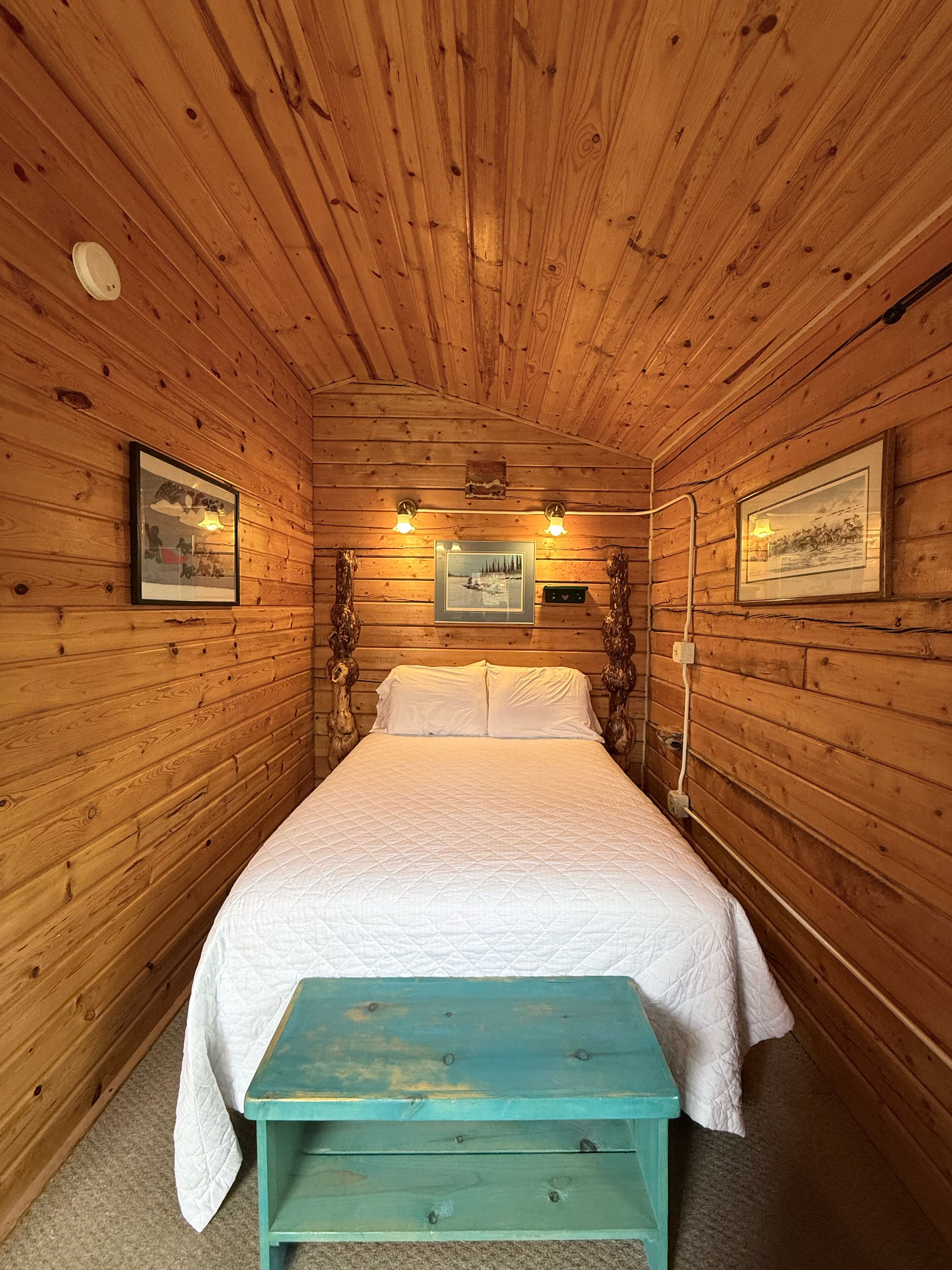 A wooden cabin bedroom with a bed, two wall-mounted lights, framed pictures, and a wooden bench.