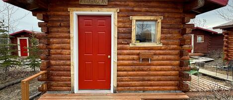 Una cabaña de troncos con puerta roja, una ventana pequeña y una terraza de madera.