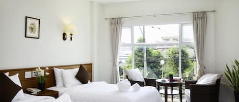A hotel room with two beds, a small table with a lamp, a wicker chair, and a view of greenery outside.