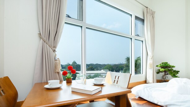 A room with a wooden table, a bed, a window with curtains, a potted plant, and a view of greenery outside. - Kanchanaburi City Hotel (Kanchanaburi)