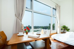 Una habitación con una mesa de madera, una cama, una ventana con cortinas, una planta en maceta y vistas al exterior con vegetación.