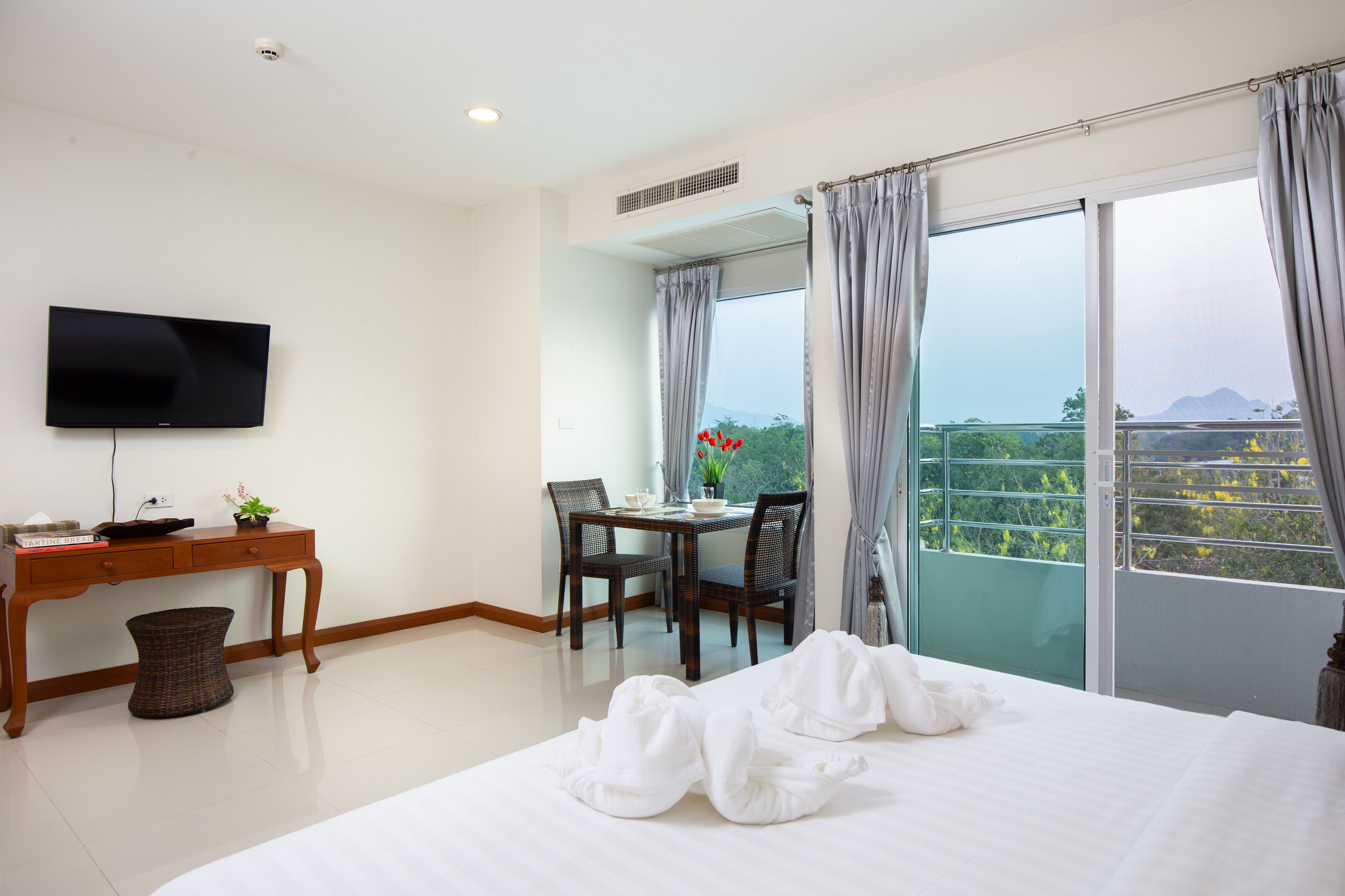 A hotel room with a bed, a desk with a TV, a dining table with chairs, and a balcony with a view.