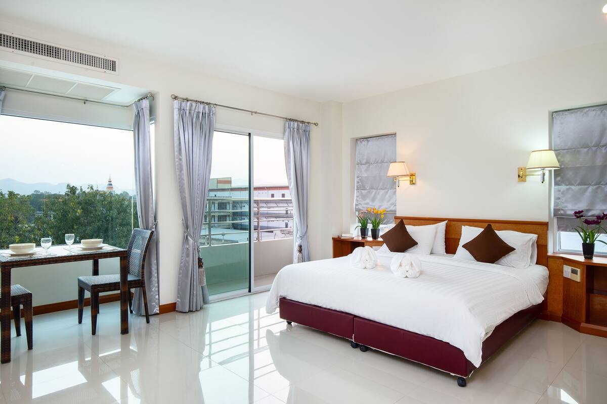 A hotel room with a bed, a desk with chairs, a view of the city, and a balcony.