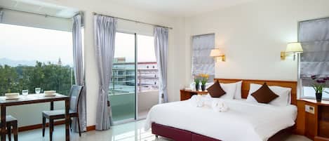 A hotel room with a bed, a desk with chairs, a view of the city, and a balcony.