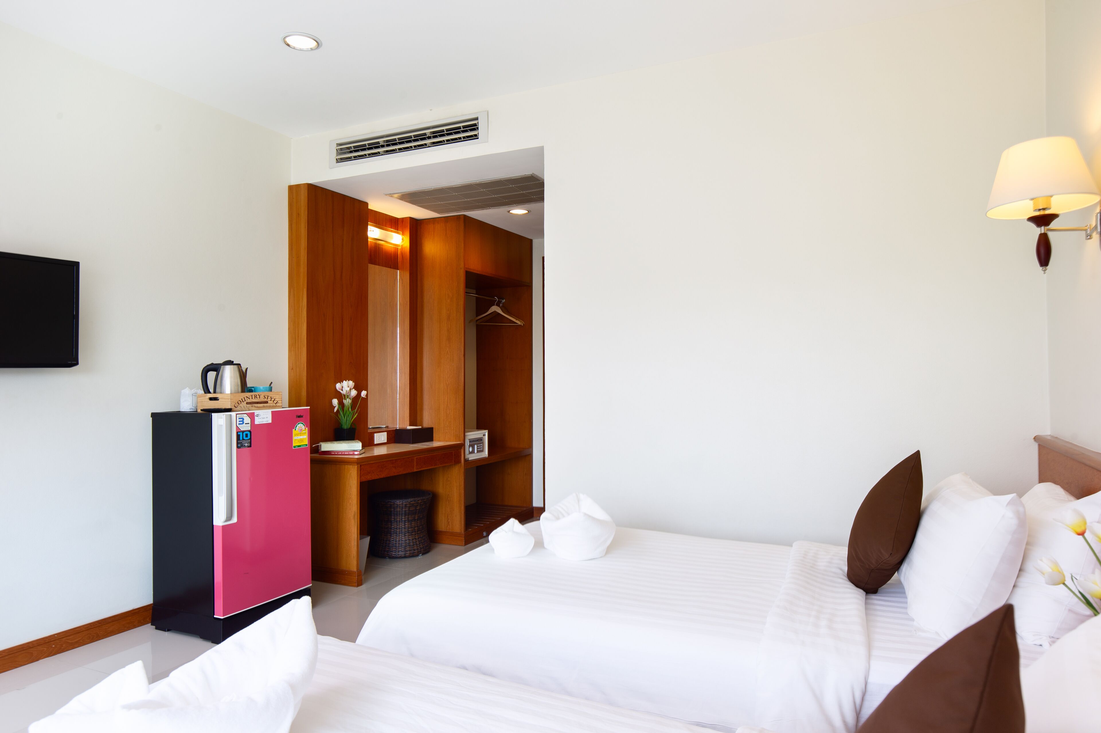 A hotel room with two beds, a wooden wardrobe, a small kitchenette, and a TV mounted on the wall.