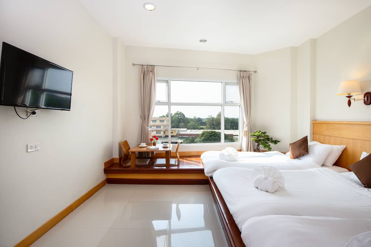 A hotel room with a bed, a TV mounted on the wall, a window with curtains, a small table with a chair, and a plant.