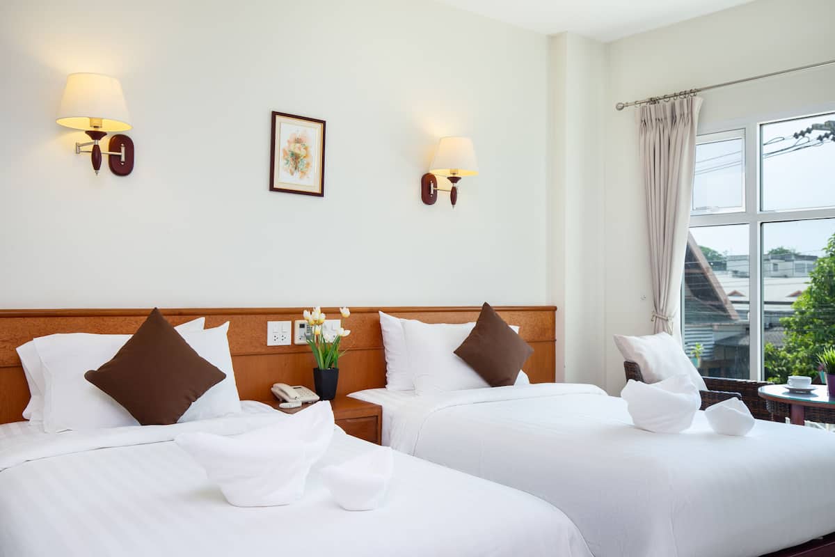 A hotel room with two beds, a wooden headboard, wall-mounted lamps, a framed picture, and a window with a view of buildings and greenery.