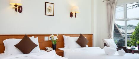 Habitación de hotel con dos camas, cabecero de madera, lámparas fijadas a la pared, un cuadro enmarcado y una ventana con vistas a edificios y vegetación.