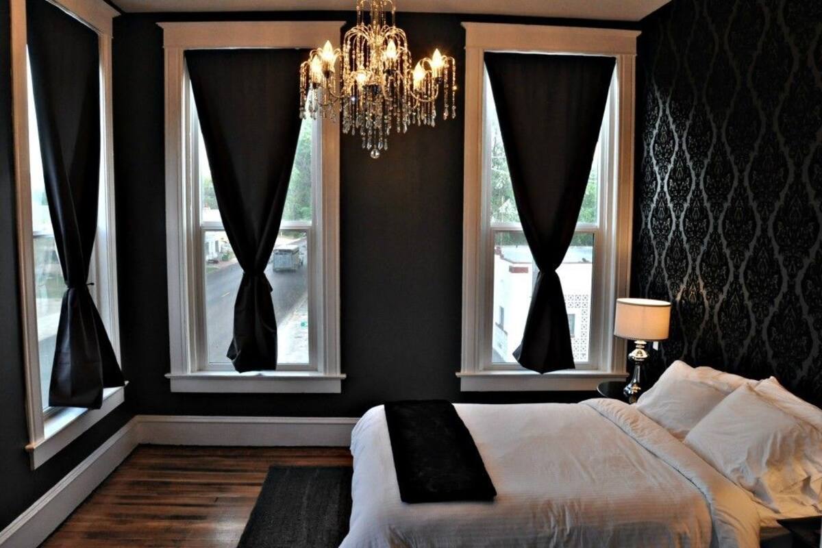 A bedroom with a large bed, a chandelier, and two windows with curtains.