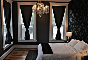 A bedroom with a large bed, a chandelier, and two windows with curtains. - The Harkness Hotel (McCammon)