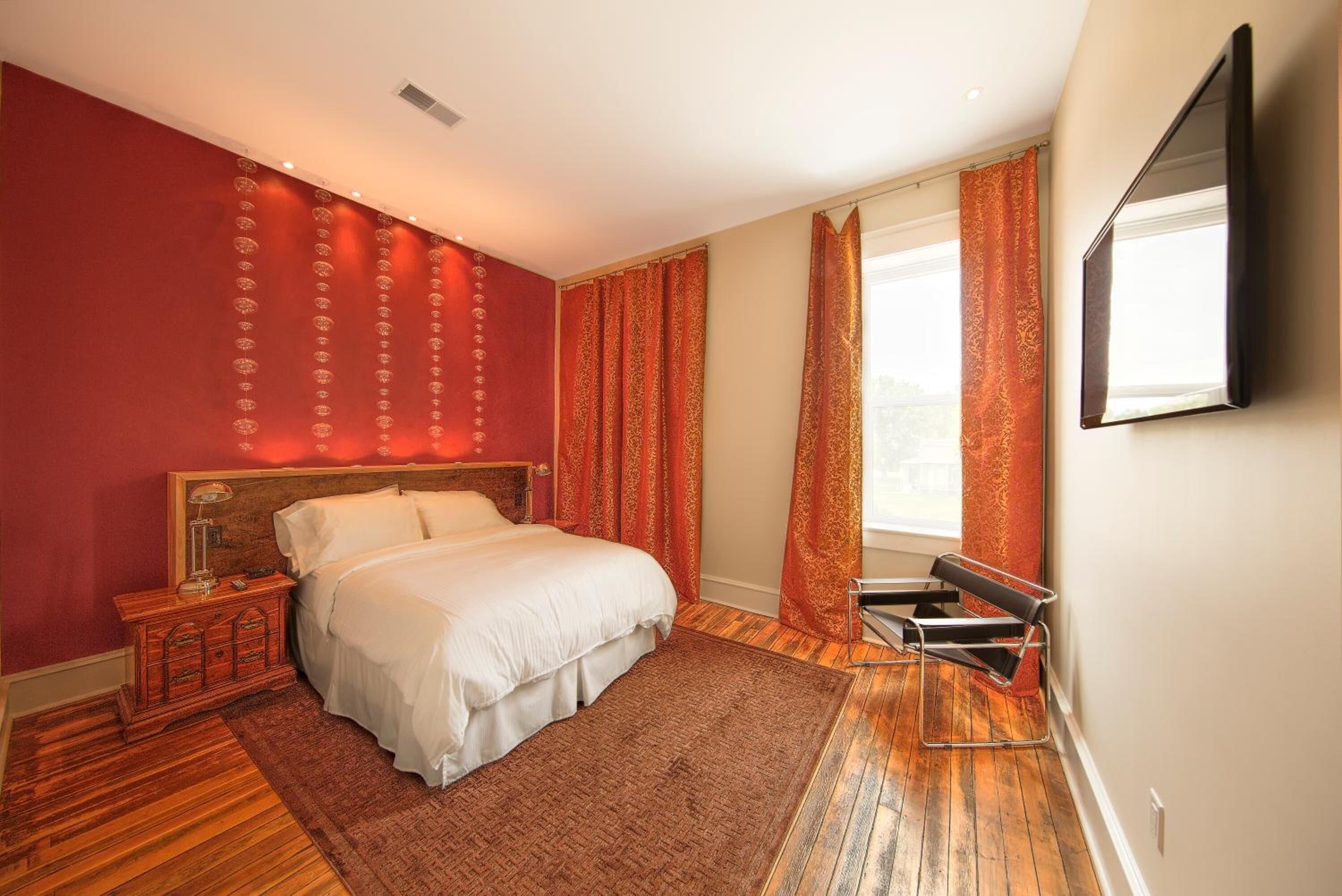 Un dormitorio con cama, suelo de madera, una mesita de noche, pared roja y ventana con cortinas.