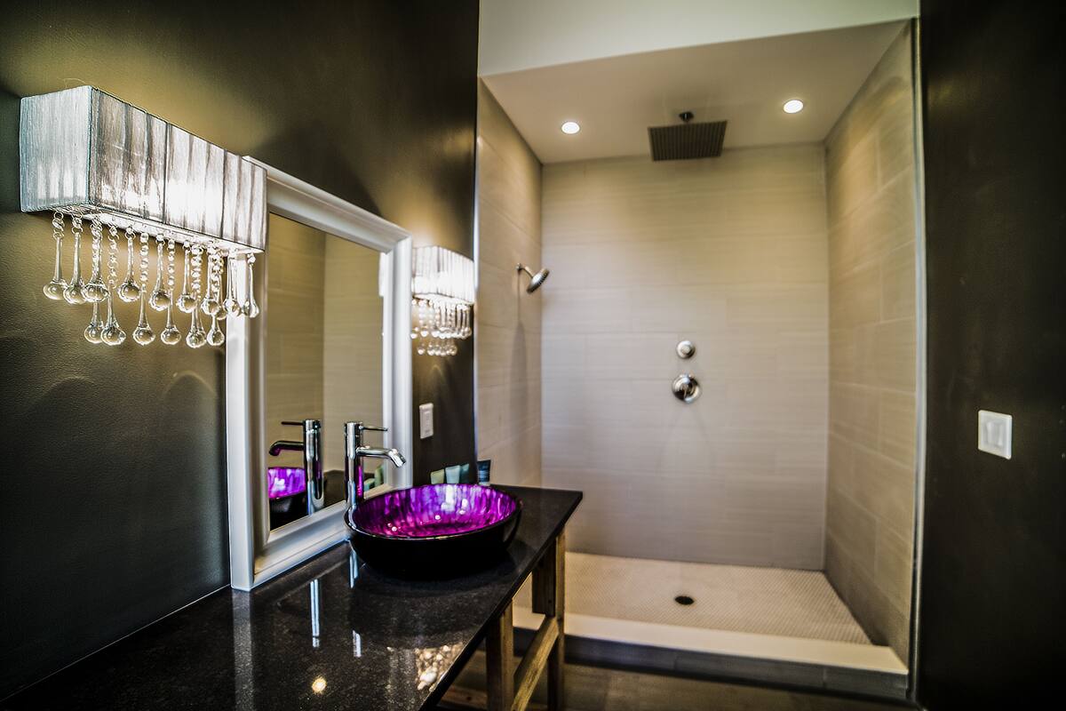 A modern bathroom with a black countertop, a purple glass bowl sink, a mirror with hanging lights, and a walk-in shower.