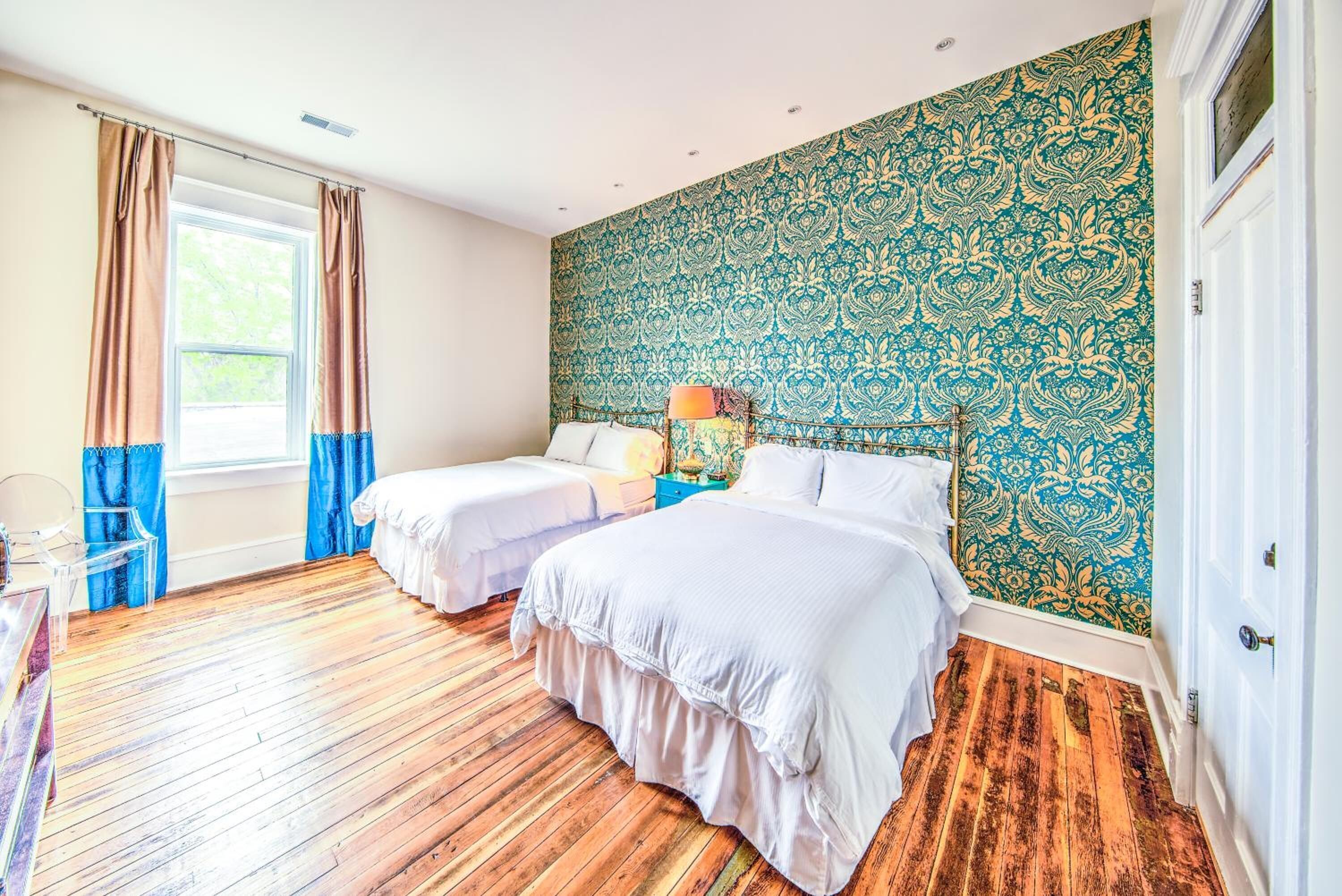 Ein Zimmer mit zwei Betten, einem fensterbelaubten Fenster, einer gemusterten Wand und einem Holzboden.