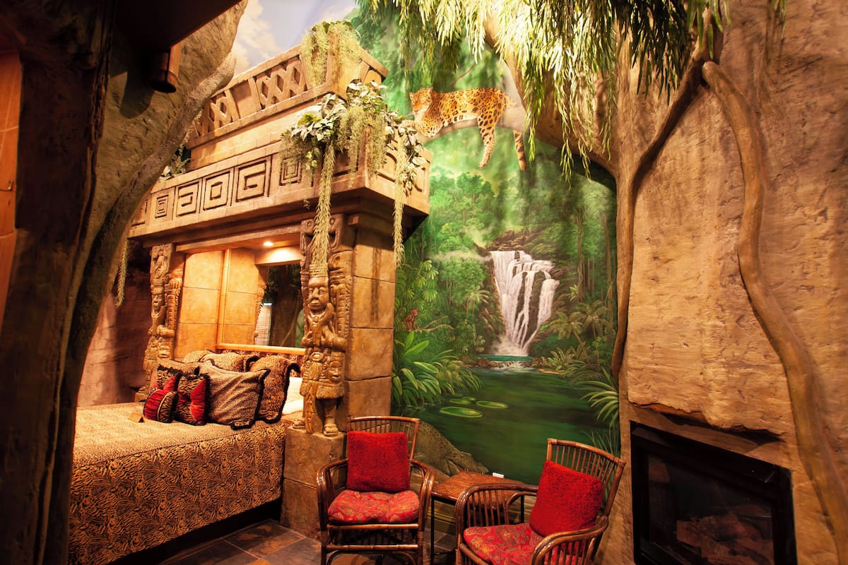 A room with a bed, two chairs, a fireplace, and a mural of a waterfall.