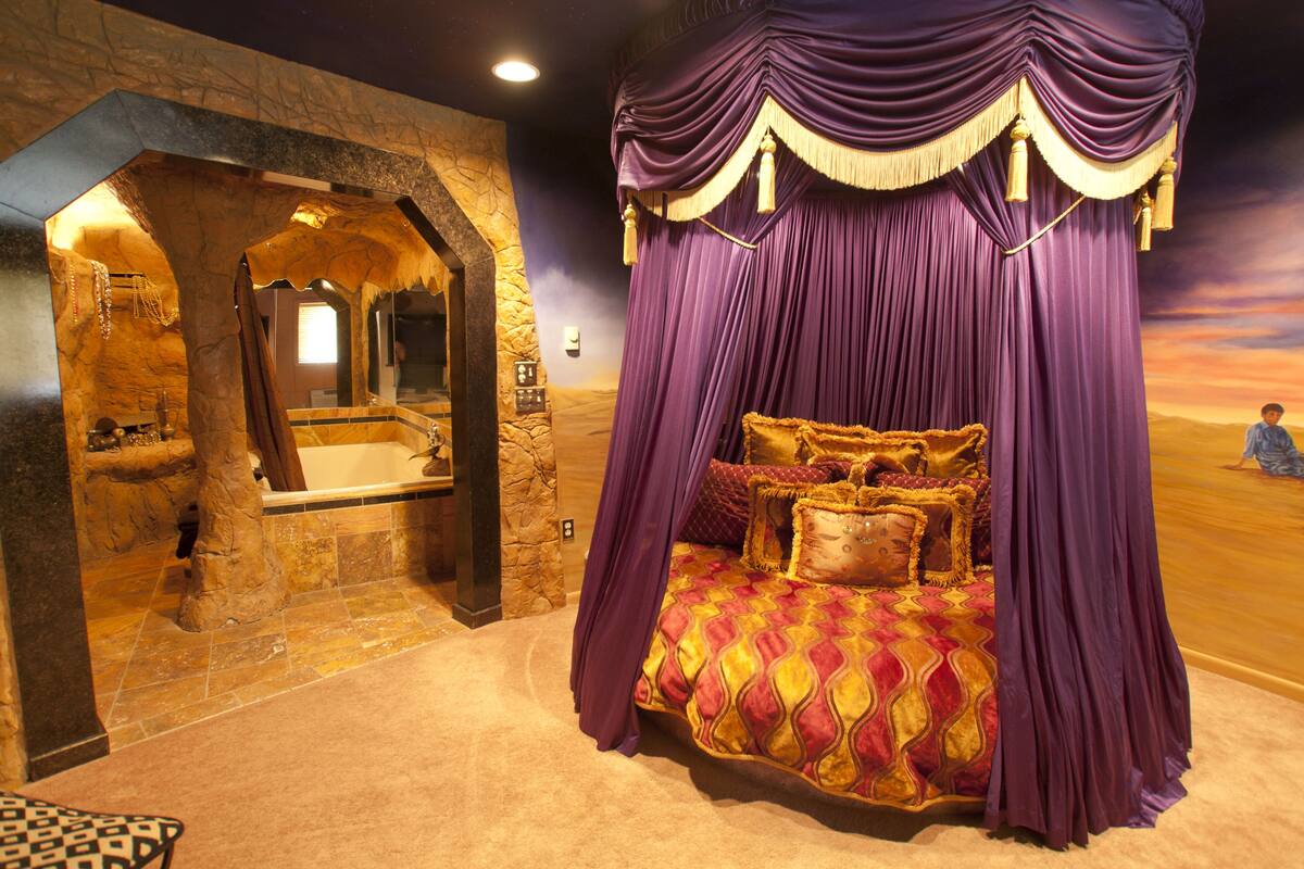 A room with a stone archway leading to a bathroom, a large bed with a purple canopy, and a desert-themed mural on the wall.