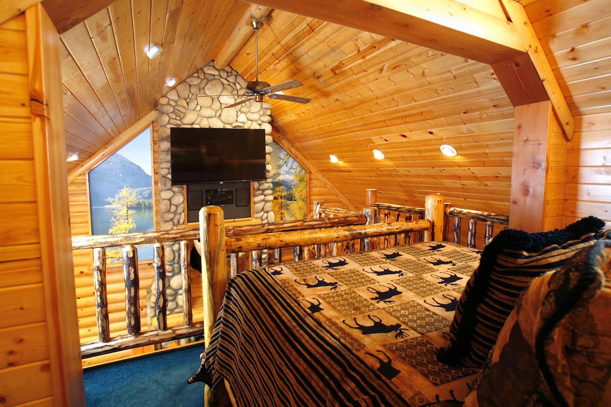 A log cabin bedroom with a stone fireplace, a ceiling fan, and a large bed with patterned bedding.