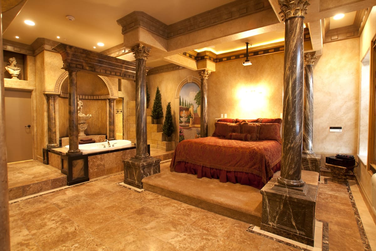 A luxurious bedroom with a large bed, marble columns, a marble bathtub, and a stone fireplace.
