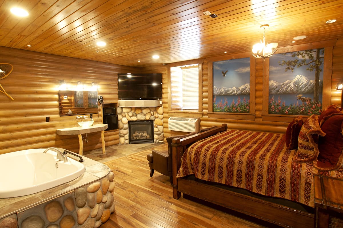 A log cabin interior with a stone sink, a large bed, a fireplace, and a painting of mountains.