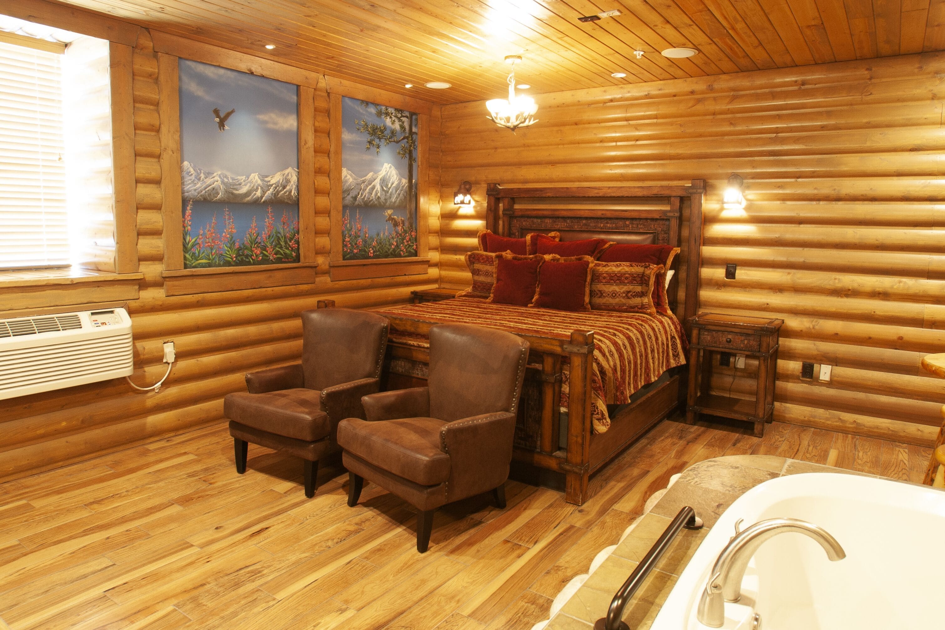 Quarto de cabana de tronco com uma cama, dois poltronas, uma mesinha de cabeceira, um lustre e um quadro de montanhas.