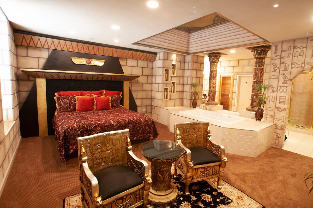 A luxurious bedroom with a large bed, a sitting area with ornate chairs, a bathtub, and Egyptian-themed decor.