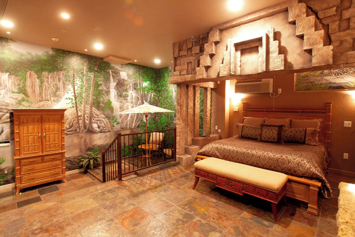 A bedroom with a stone wall, a bed with multiple pillows, a wooden bench, and a small table with an umbrella.