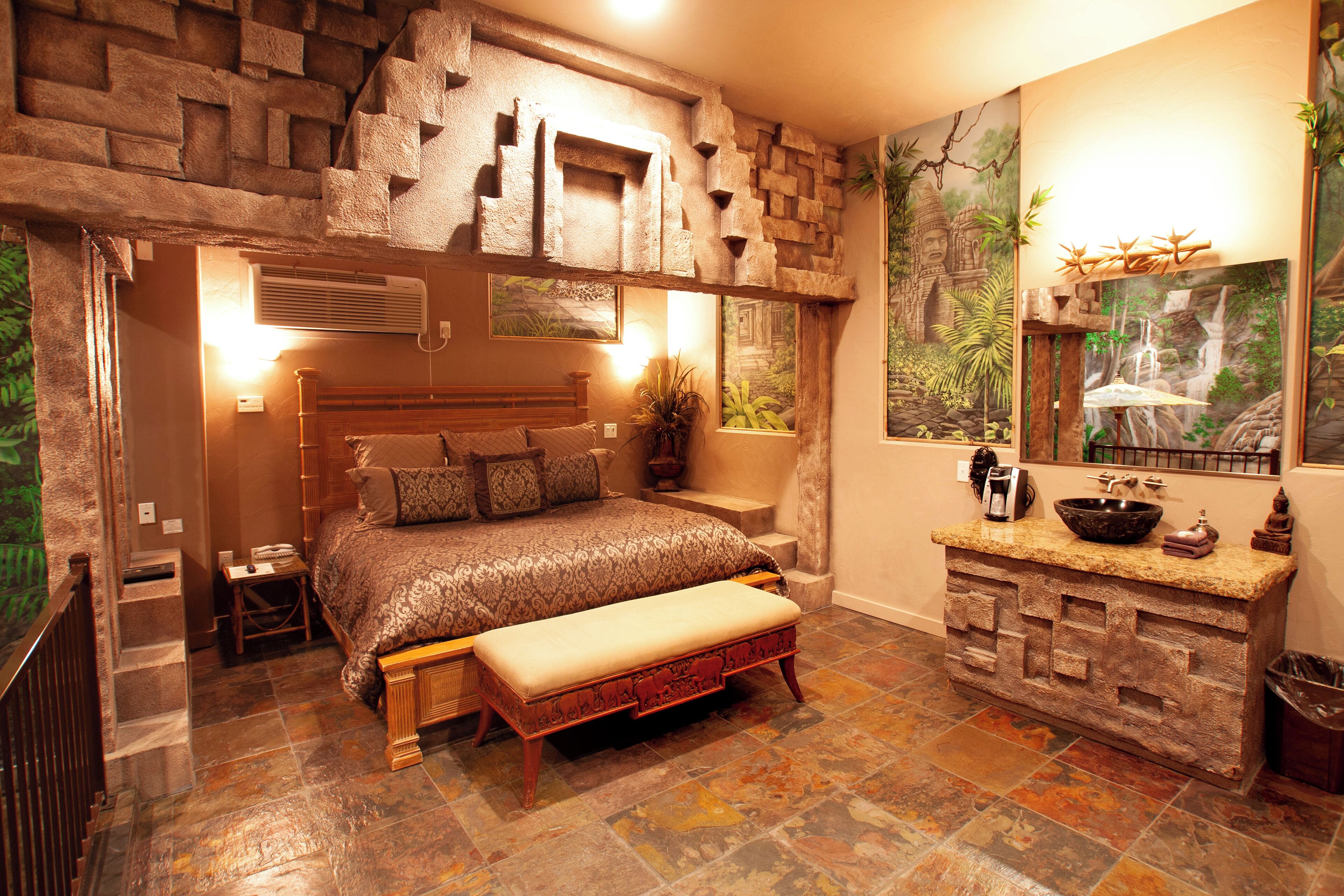 A bedroom with a stone wall, a bed with a headboard, a bench, a window with a view of a waterfall, and a sink with a stone countertop.