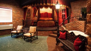 A cozy room with a brick wall, a bed with red bedding, a staircase, and a seating area with chairs and a bench. - Destinations Inn (Idaho Falls)
