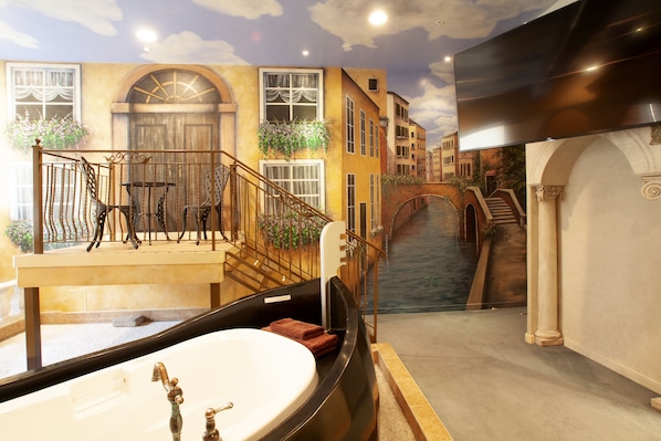 A luxurious bathroom with a large bathtub, a wooden bench, and a view of a canal with buildings. - Destinations Inn (Idaho Falls)