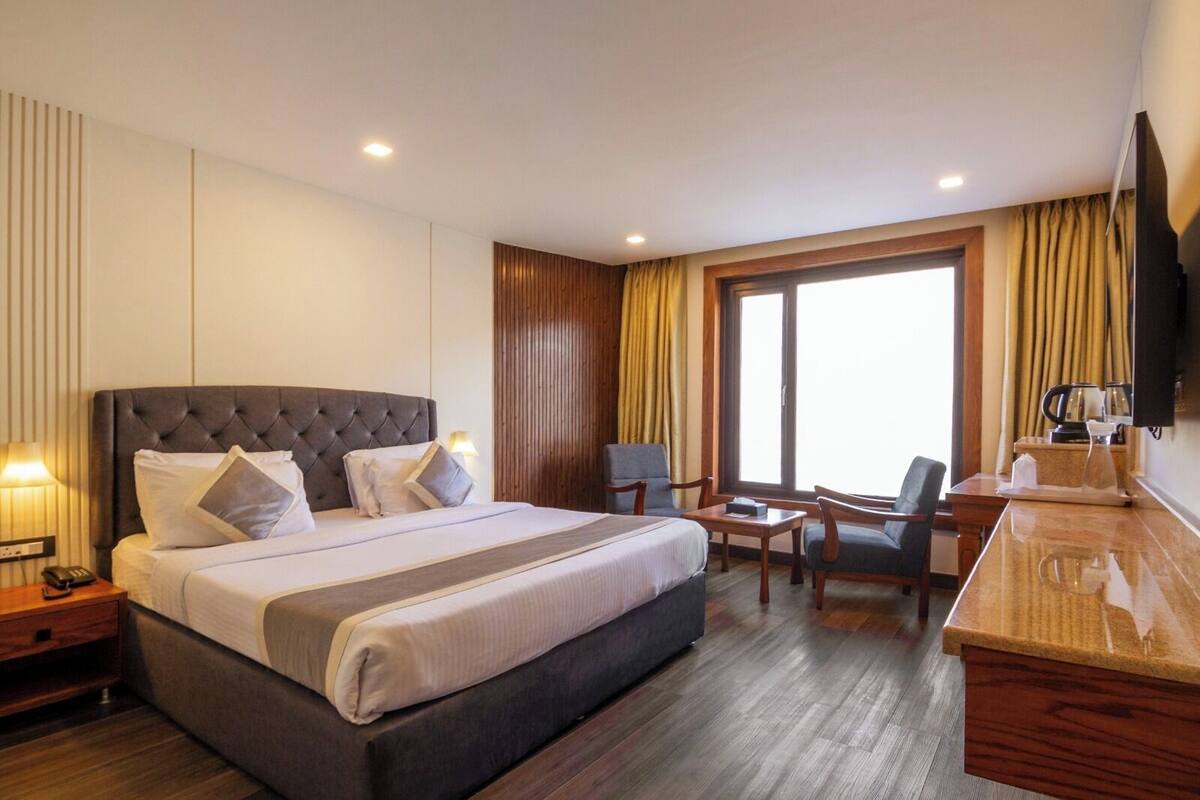 A hotel room with a large bed, bedside tables, a desk with chairs, a television, and a large window with curtains.