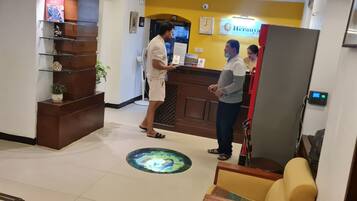 Zona de recepción de un hotel, con mostrador de recepción, un reloj digital y un cartel del hotel.