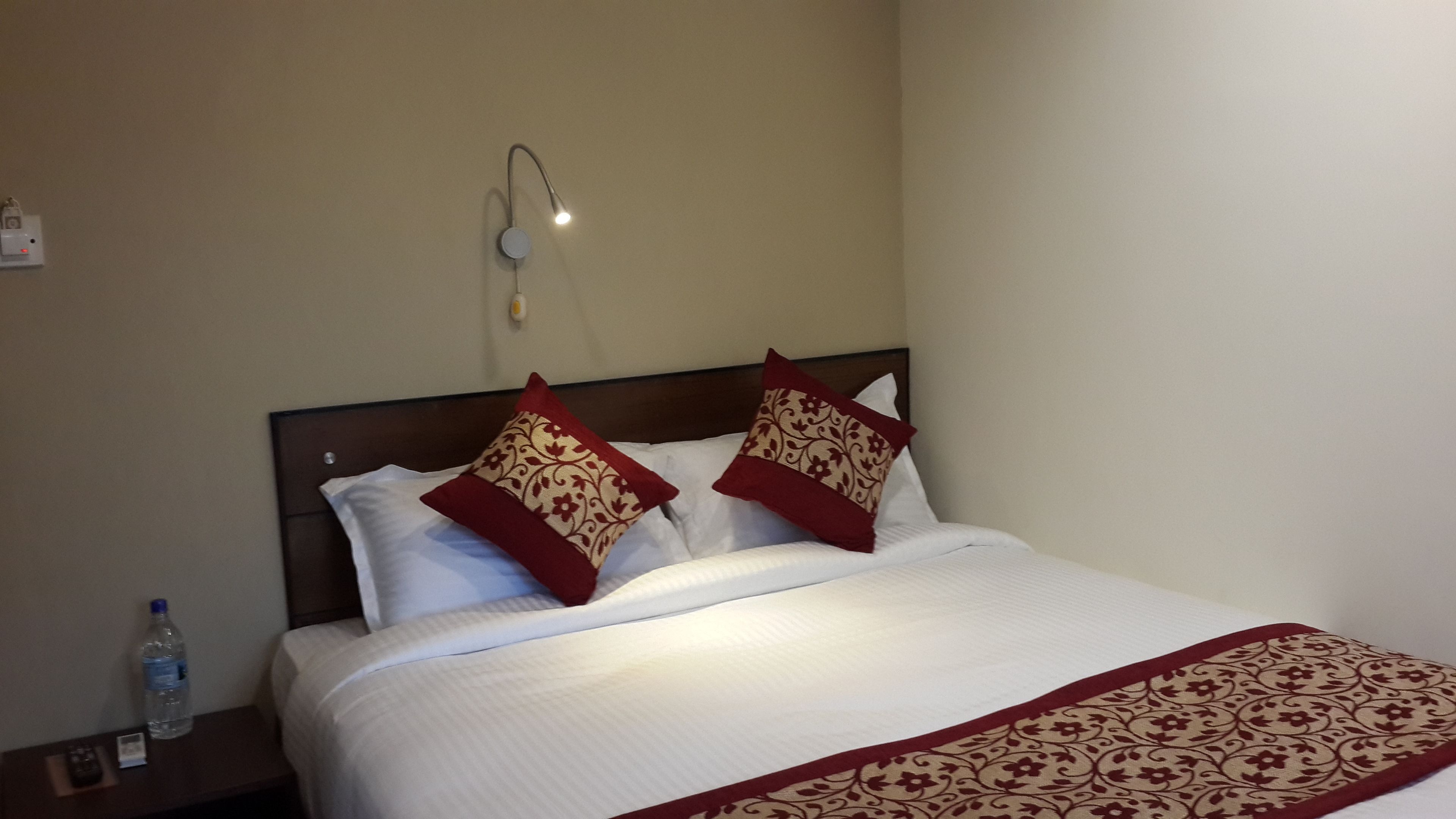 A neatly made bed with white linens and two decorative pillows, a bedside table with a bottle of water, and a wall-mounted light fixture.