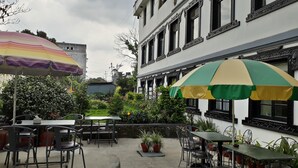 Un área de café al aire libre con mesas y sillas, rodeada de macetas con plantas y un edificio con marcos de ventanas ornamentados.