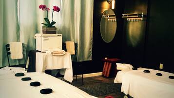 A hotel room with two beds, a small table with a lamp, a mirror, and a window with curtains.
