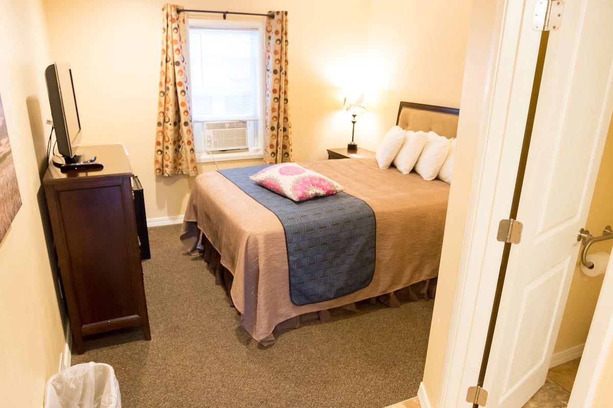 A bedroom with a bed, bedside table, lamp, window with curtains, and a door.