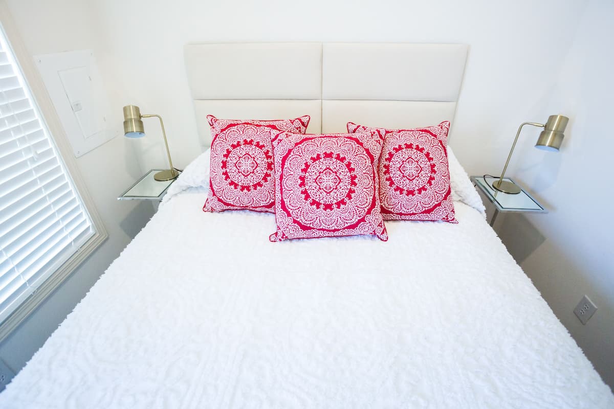 A neatly made bed with white bedding and three decorative pillows, two bedside tables with lamps, and a window with blinds.