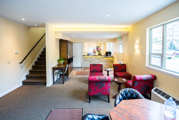 A hotel lobby with a staircase, a reception desk, a kitchen area, and a seating area with floral-patterned chairs.