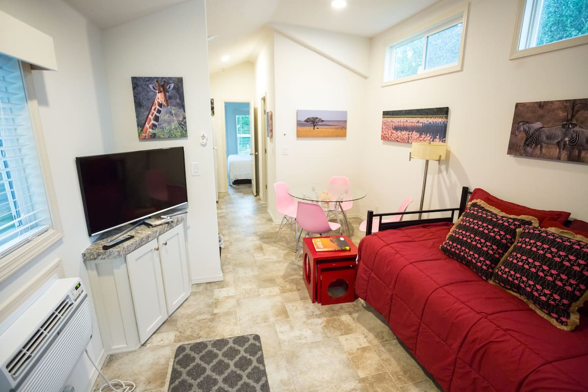 A compact living space with a television, a red sofa, a small table, and a chair. There are paintings on the walls and a window with blinds.