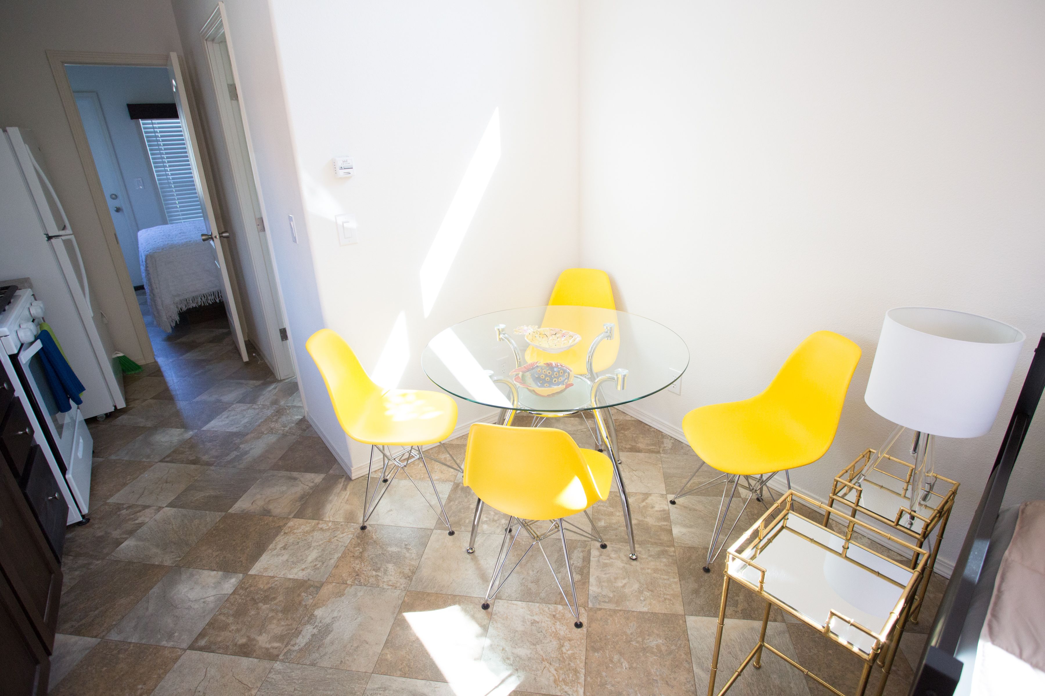 Un coin repas avec une table en verre et quatre chaises jaunes, une lampe blanche, et un espace cuisine avec des placards et un réfrigérateur.