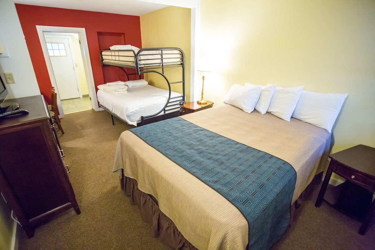 A hotel room with a bunk bed, a desk with a computer, and a bedside table with a lamp.
