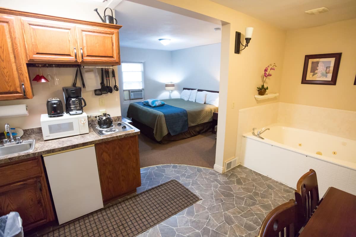 A compact kitchen with a microwave, coffee maker, and sink. A bedroom with a bed and nightstand is visible through an open doorway. A bathtub is in the adjacent room.