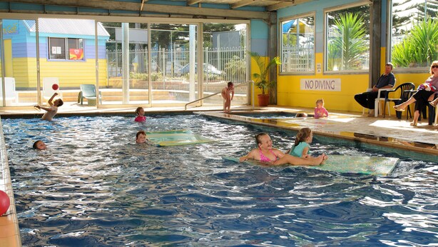Una piscina con gente en el agua y en el borde, un cartel de 'NO DIVING' y un edificio con un letrero de 'Caf' al fondo.