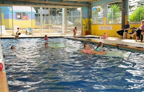 Una piscina con gente en el agua y en el borde, un cartel de 'NO DIVING' y un edificio con un letrero de 'Caf' al fondo.