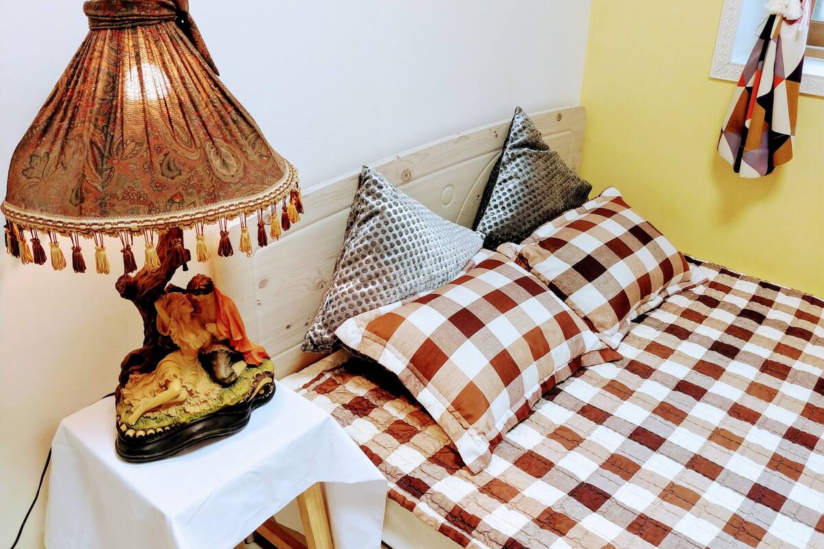 Una cama con colcha a cuadros, una lámpara con pantalla estampada y almohadones decorativos sobre una mesita de noche blanca.
