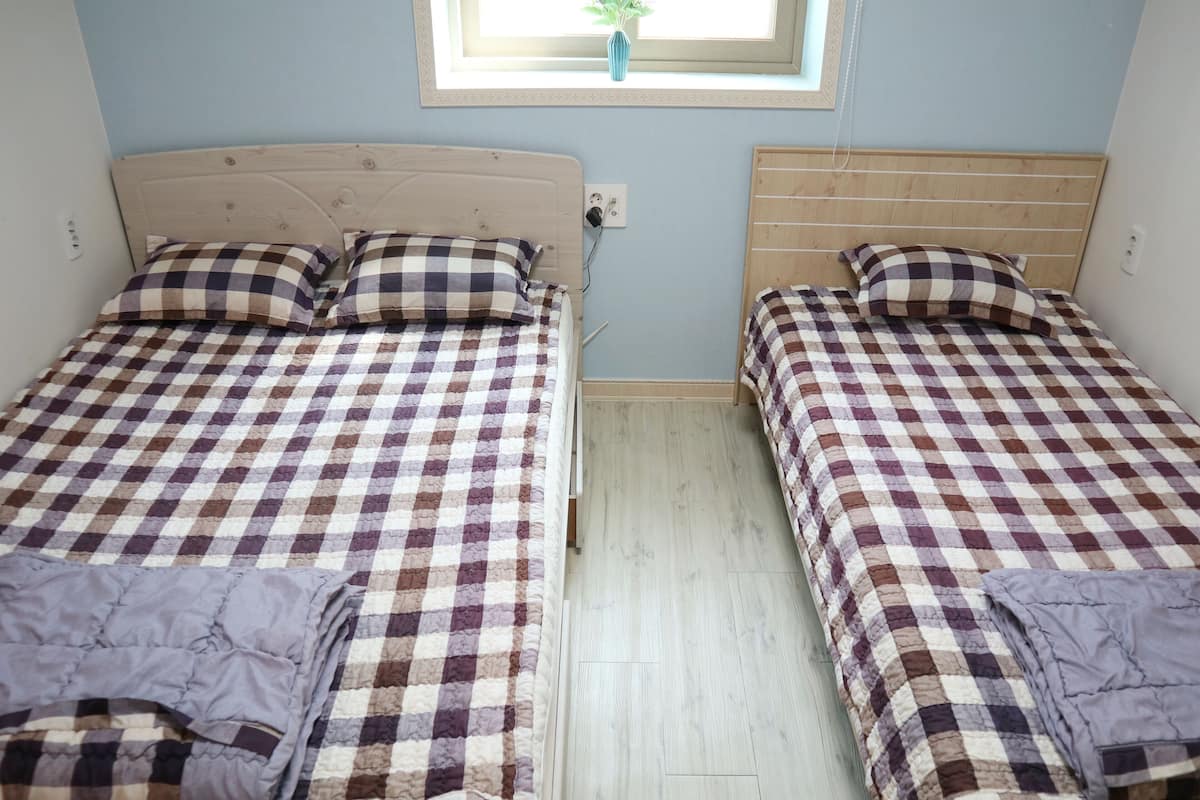 Dos camas individuales con ropa de cama a cuadros, un cabecero de madera, una ventana pequeña con una planta y un interruptor de luz en la pared.