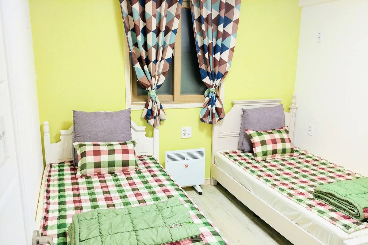 Una habitación con dos camas, cada una con una colcha y almohadas a cuadros, una ventana con cortinas estampadas y un calentador fijado a la pared.