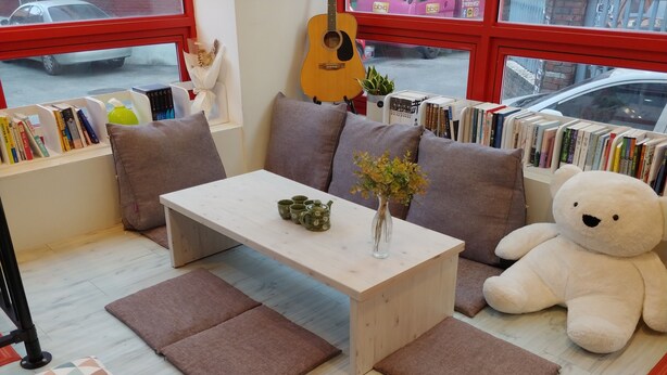 A cozy reading nook with a wooden table, cushions, a large teddy bear, and a bookshelf filled with books.