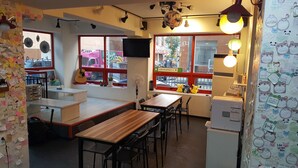 A cozy cafe with wooden tables and chairs, a large window with a view of the street, and various decorations including notes and stuffed animals.