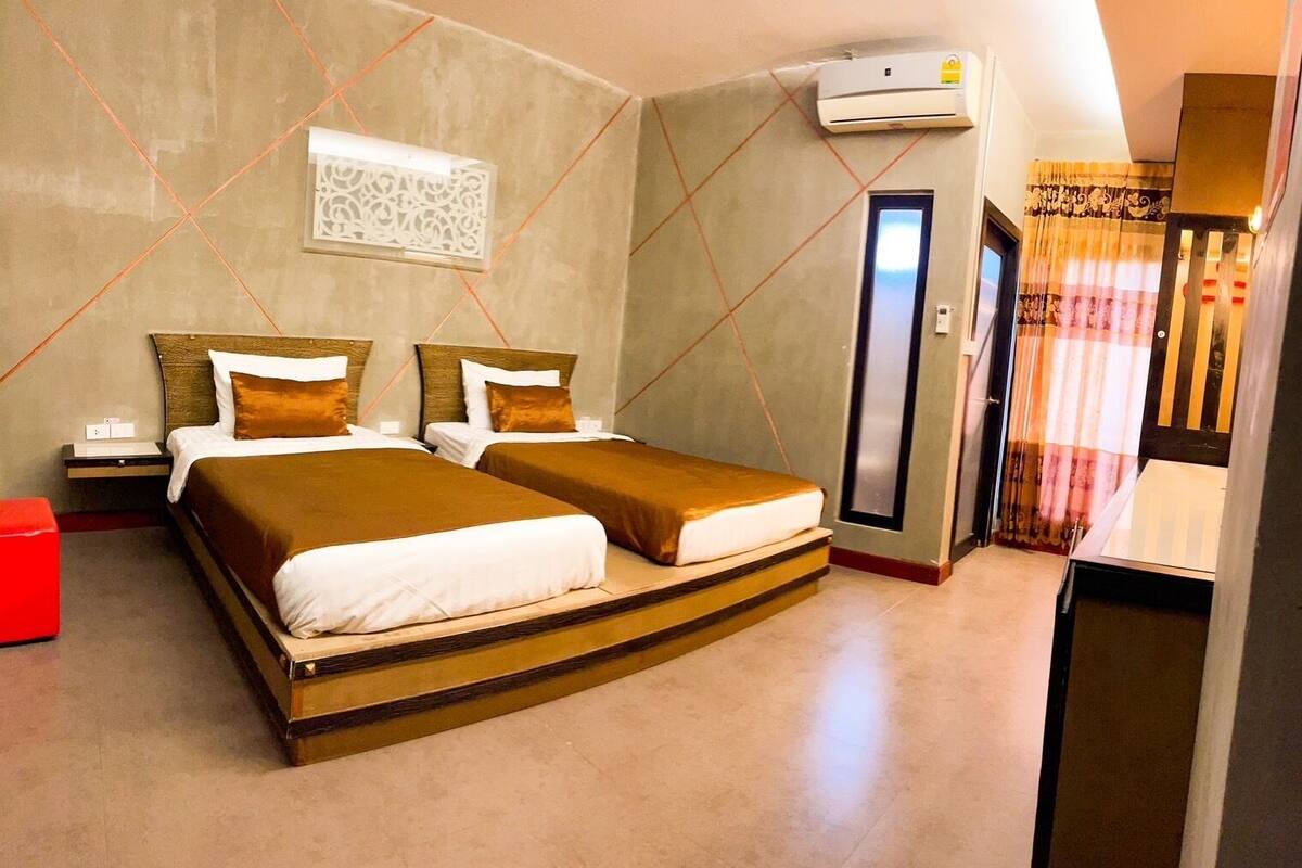 Habitación de hotel con dos camas, un bote de basura rojo, un equipo de aire acondicionado, una puerta y una cortina.