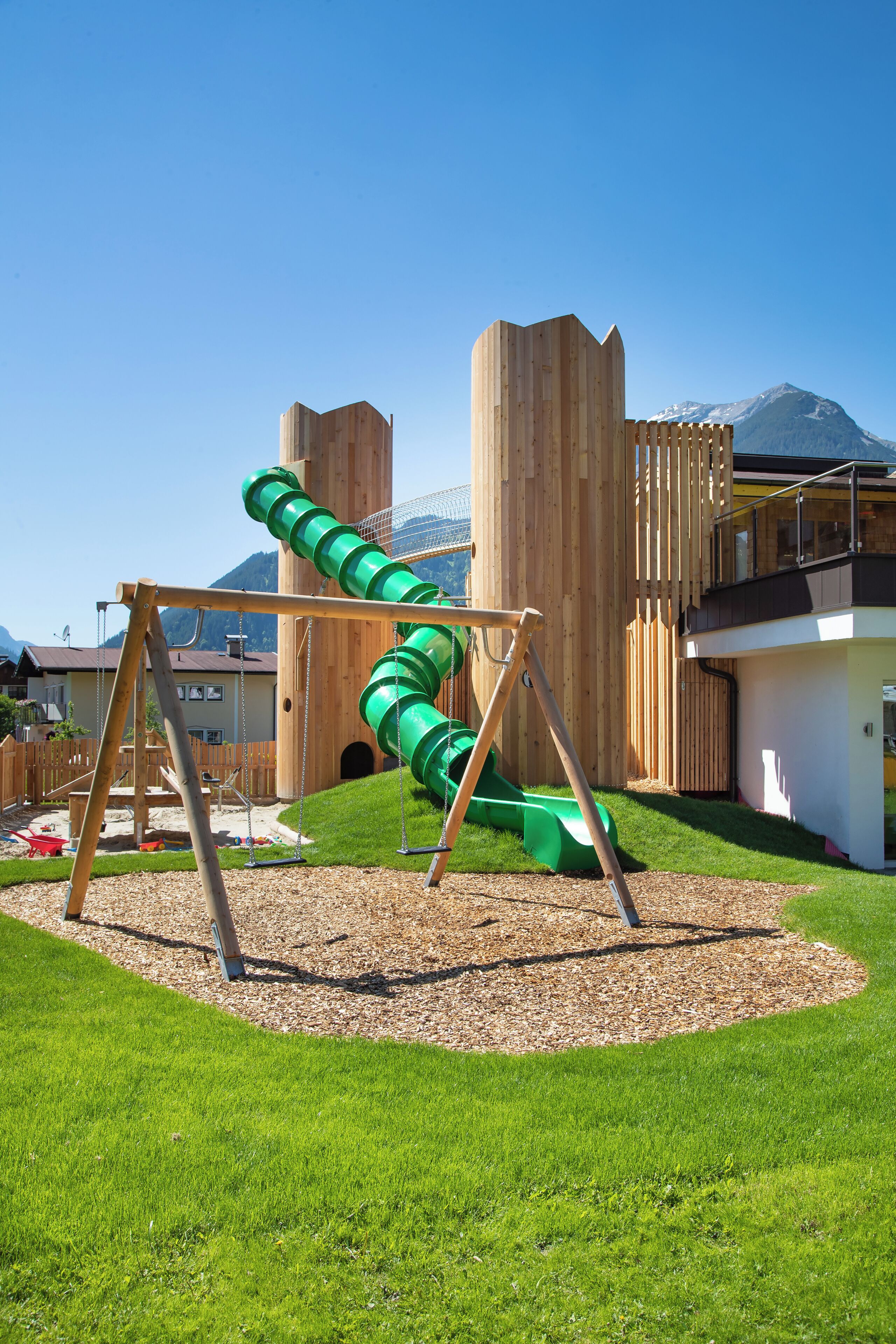 Un'area giochi con uno scivolo verde e altalene, circondata da un prato e strutture in legno.